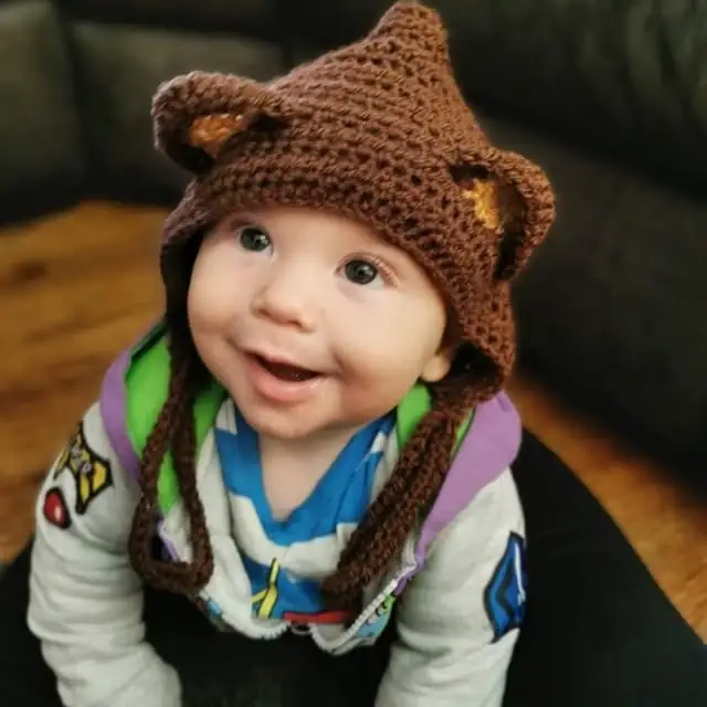 A smiling baby in a brown crochet bear bonnet with little ears