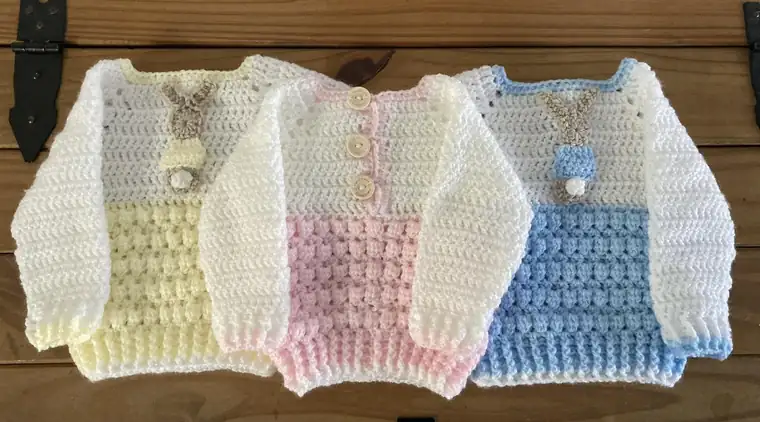 Three handmade crochet baby jumpers in lemon, pink and blue, two with a little bunny on the front