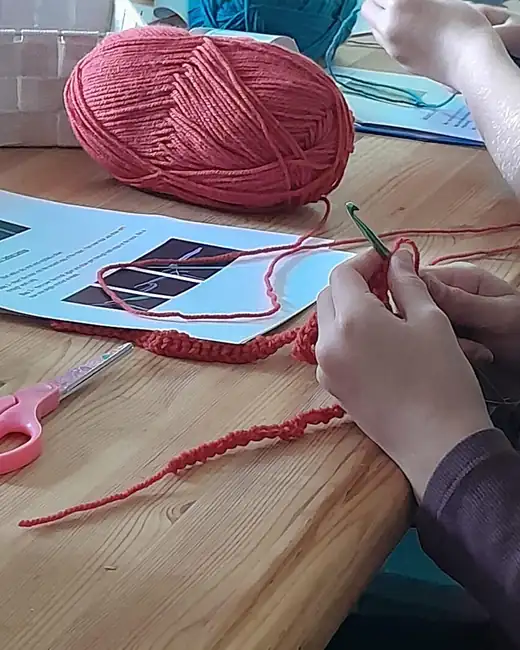 Hands working a crochet chain in red yarn next to a printed stitch guide