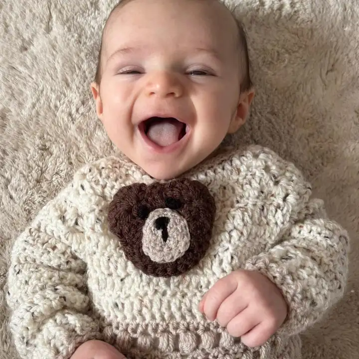 A laughing baby wearing a cream crochet jumper with a brown teddy bear face